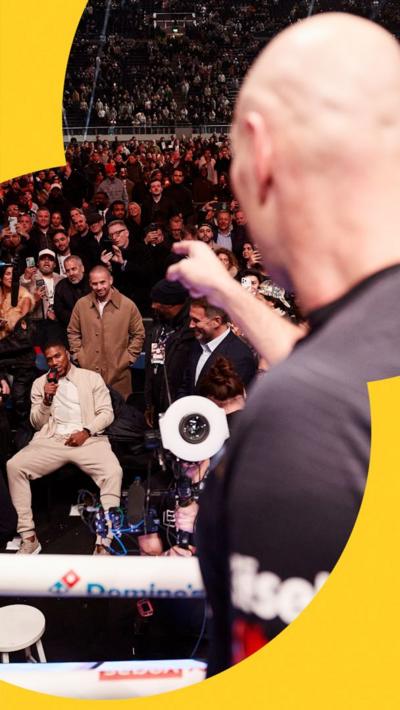 Tyson Fury pointing at Anthony Joshua. Fury is wearing a black shirt, and Joshua a cream jacket.