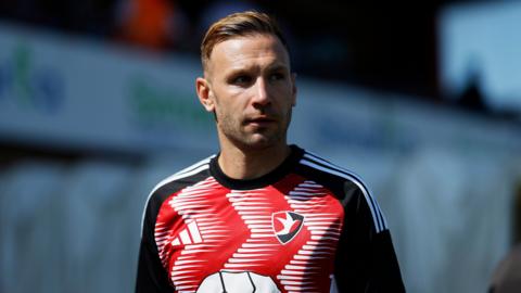 Andi Weimann standing on the pitch during pre-match training wearing a red and white Cheltenham shirt with black sleeves.