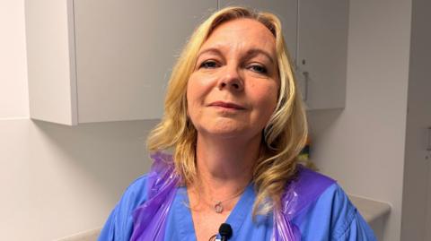 A head shot of a female doctor, in blue scrubs, wearing a plastic apron, standing in an examination room