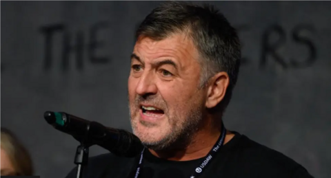 A man with short, black hair and stubble stands in front of a microphone mid-speech. He is wearing a black t-shirt and has a lanyard around his neck