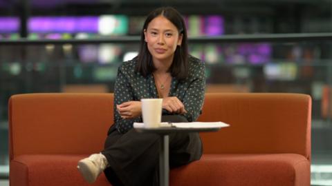 A woman is sat on an orange sofa wearing a green patterned shirt, dark trousers and white shoes. In the background is a blurred image of a news gallery with various screens. There is also a small table in front of the woman with a white mug and paper. 
