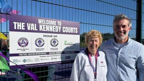 A woman wearing a white jumper and purple lanyard with short curly blonde hair stands beside a man in a blue shirt with short grey hair and beard. They are stood in front of a sports court with a sign that reads: Welcome to The Val Kennedy Courts.