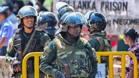 A member of the army stands in front of a yellow barrier - a building with the words Sri Lanka prison is in the background. Four other personnel are behind him, one facing the same way 