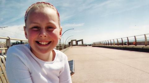 A young girl with a white top sits at a beach promenade