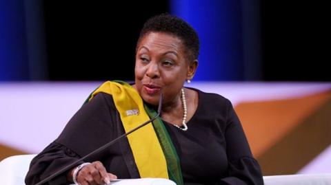 Olivia Grange, a Jamaican woman with short black hair, is sitting down in front of a microphone. She is wearing black clothes and a black, yellow and green sash. She is wearing a pearl necklace and the background is blurred. 