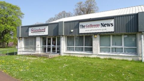 An industrial building with signs for the Dumfries and Galloway Standard and the Galloway News on its front