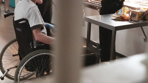 Inmate in a wheelchair. You cannot see the man's head. He is in a room behind bars where food is being served 