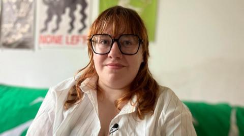 A woman with auburn hair and large black glasses looking at the camera. She's wearing a white pinstriped shirt sitting on a sofa with a green drape on the back, with other posters behind on the wall. 