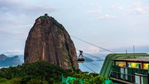 A view of Sugar Loaf cable car at Guanabara Bay in November 2024. The steep rocky mountain can be seen against the blue of the bay. At the top of the mountain, the cable car station is visible.  Cables connect it with Morro da Urca, from which the photo is taken. A cable car can be seen suspended halfway between the two. 