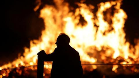 A man stands near a bonfire at Ottery St Mary on Guy Fawkes night in Britain. He is silhouetted against the orange flames.