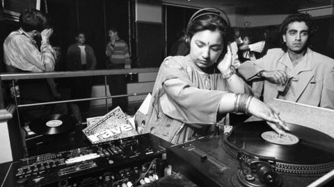 A black and white photo of a young woman spinning a record on DJ decks. She is wearing traditional South Asian clothing as well as a bindi and bangles. Groups of young people stand or dance in the background.