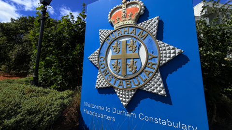 A close up of a big blue sign with Durham Constabulary's crest is in front of the white Durham Police headquarters. A tiny part of the building can only just be seen beyond thick green hedges. The sign stands in a grassy park area with a traditional police lamppost in the background. 