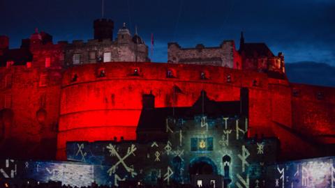 Edinburgh Castle is lit up in red lights. the front gate is lit up in a blue light with white markings that resemble cave markings.