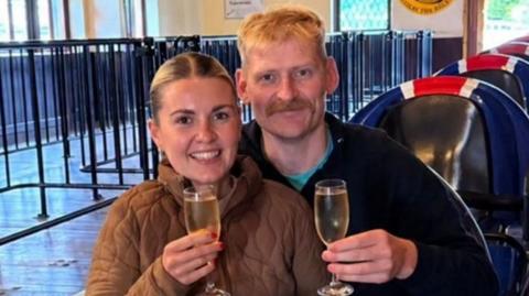 Nicolle and Derek sit together in an Avalanche rollercoaster car at Blackpool Pleasure Beach, holding glasses of sparkling wine in the ride station