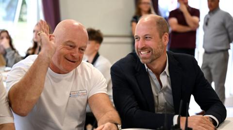 Ross Kemp waves one hand in his air as he smiles. He is sat next to Prince William who laughs as he looks at Kemp.