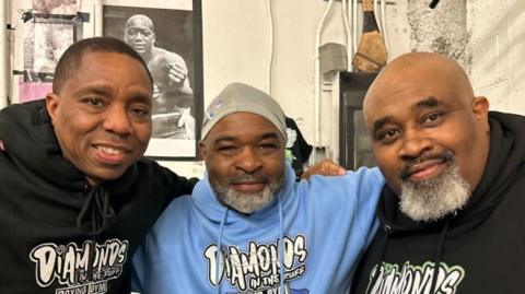 Three men stand with their arms around each other. They are wearing matching branded hoodies in black and blue. In the background is a black and white image of a boxer.