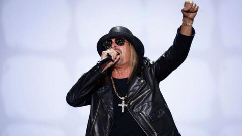 Musician Kid Rock sings into a microphone and lifts a fist. He is wearing a black leather jacket, T-shirt, hat and sunglasses and a large silver cross on a chain.