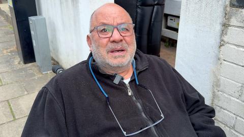 Yehia Hassan, who is wearing glasses and is in a wheelchair outside his house, has an anguished expression  