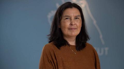 A woman wearing a brown top stands against a blue background