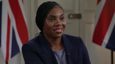 A head and shoulders picture of Kemi Badenoch wearing a navy suit with a grey shirt. She is sitting in front of two British flags.