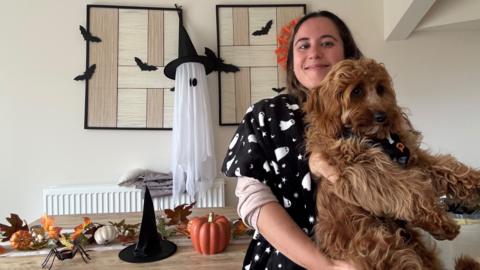 A young woman shows off her spooky Halloween decorations in her living room as she holds her puppy and smiles. She is wearing a black scarf with drawings of white ghosts. Behind her, a table with a witch's hat and pumpkin decorations, a fake spider, and hung on the wall is a ghost wearing a witch's hat surrounded by bats.