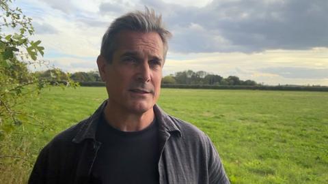 Michael Ergatoudis is standing in a green field with tree in the distance.  He has greying, medium length hair and is wearing a dark shirt over a black t-shirt.