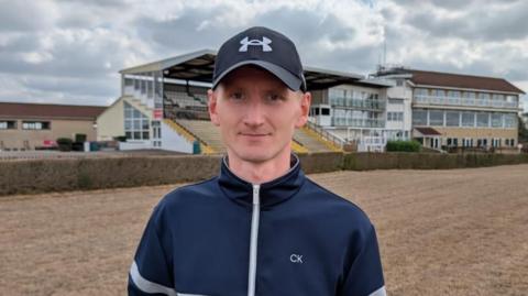 Tom Ryall, a man wearing a navy tracksuit top and baseball cap, is standing on a horse racing track with buildings in the background. The ground is visibly brown. 