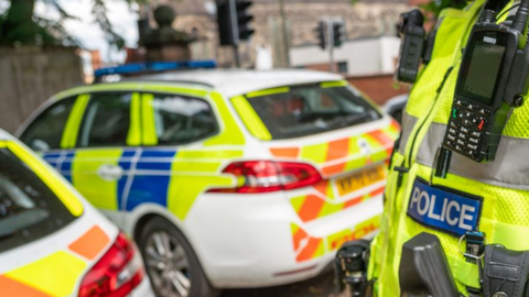 Close up of the Police badge on the front of a hi-vis jacket, with two police cars in the background