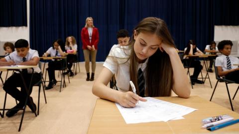 A female secondary school student sits an exam in a spacious school hall, the atmosphere calm and focused. She concentrates intently on her paper, pen in hand. The mid shot captures her quiet determination in the academic setting.