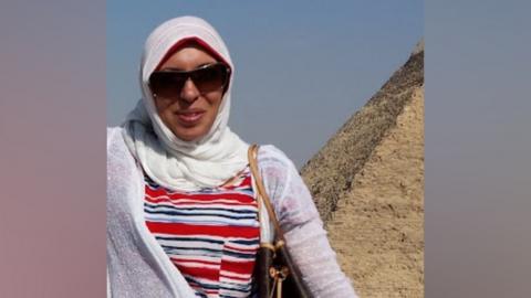 A woman in a white headscarf, striped shirt and white cardigan smiles at the camera. She also wears black sunglasses. She stands next to a pyramid.