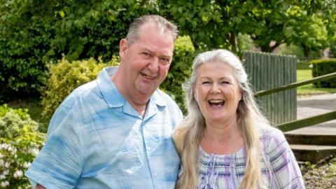 Tom and Jayne are stood in a park or garden area. Tom is wearing a blue short-sleeved shirt and Jayne is wearing a striped dress in lavender, purple and white.