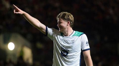 Freddie Draper looks to his right as he holds his right arm up into the air with his finger pointed after scoring a goal. He is wearing white Lincoln City kit
