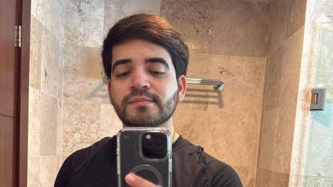 César Gastélum looks into a mirror as he takes a selfie. He has a beard and black hair. He is wearing a black T-shirt and a gold chain around his neck 