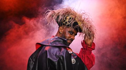 An actor dressed as pantomime pirate is presented against a red backdrop. He is wearing a black outfit with a red shirt as well as a wide-brimmed black hat with a plume of orange feathers on top.