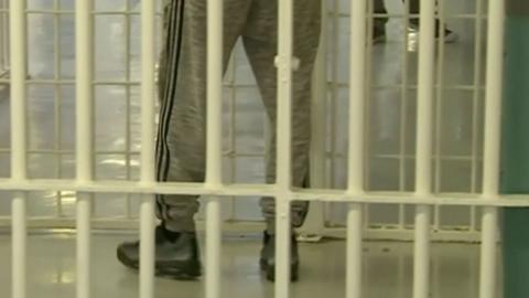 An image shows yellow painted prison bars with a person standing behind them with only their legs visible. They are wearing light grey tracksuit trousers with a double stripe down the side of the legs in a darker colour. They are also wearing grey and black trainers.