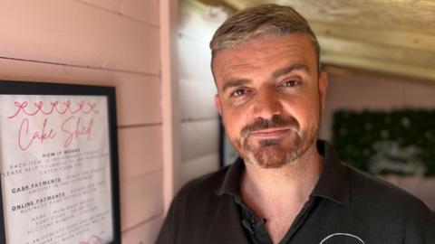 Lee McCann - a man with short light coloured hair combed over to the left - looks at the camera.
He has a worn expression on his face. He is standing in a small shed, with wooden walls painted pink.