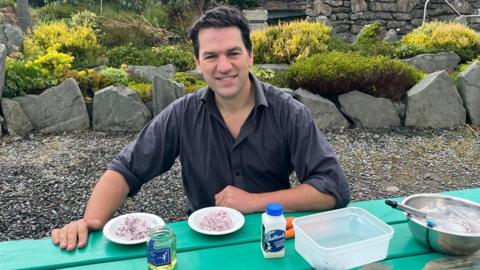 A man with dark hair, wearing a dark blue shirt, sits at a green outdoor table. He is surrounded by shrubs in a rockery. He has samples of honey and milk on the table. 