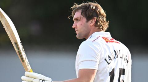 Tom Lammonby raises his bat in his left hand with his helmet off and in his right hand after passing 100 for Somerset