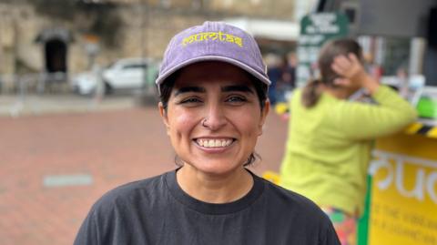 A female market trader wearing a black t-shirt and purple cap smiles at the camera