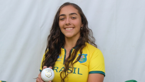 Laura Cardoso holding a white ball while wearing a Brazil cricket kit