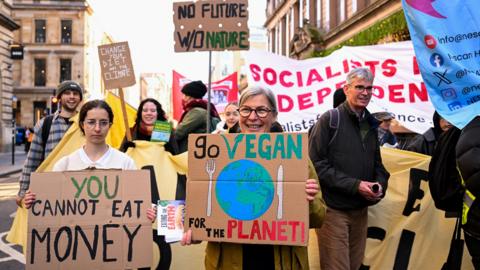 A group of people holding colourful placards at a climate march. The left placard reads 'You cannot eat money'. The middle reads 'Go Vegan for the planet'. There are several other signs in the background. The woman front centre is smiling at the camera.