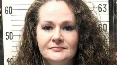 A woman with curly brown hair having a mug shot taken.