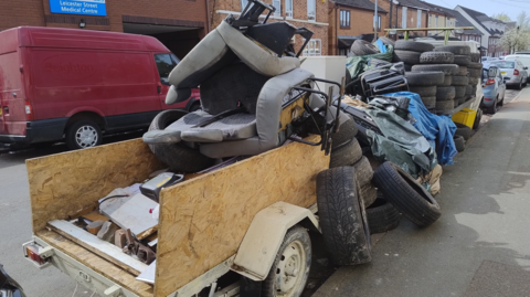 A chair, rubble and tyres are seen on the back of the vehicles with tyres stacked up. Cars are parked in the street.