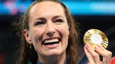 A headshot show Tatjana Smith smiling broadly as she holds up her 100m breaststroke gold medal during the medal ceremony at the Paris 2024 Olympic Games