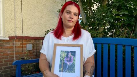 Natalie James, who has dyed red hair, is sitting on a blue garden bench. She is wearing a white T shirt and blue jeans and is holding in her lap a framed photograph of her teenage daughter sitting down on the grass.