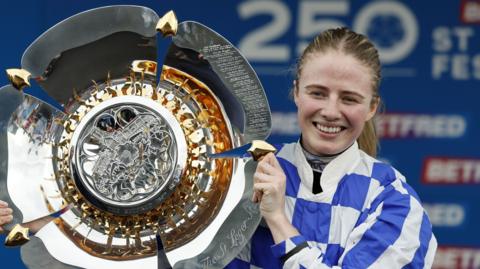 Saffie Osborne, in blue and white-checked silks, holding a silver trophy to her right
