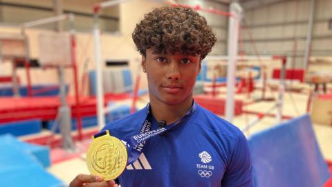 Uzair Chowdhury looking at the camera while clutching his gold medal. He is wearing his blue Team GB uniform and is stood at his gym, with crash mats and equipment blurred out behind. Chowdhury has curly brown hair and is wearing stud earrings.