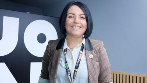 Becky Bradshaw, standing in a room, by a sign, wearing a white shirt, checked jacket, with a lanyard round her neck. She has short bobbed hair, she is smiling and looking straight at the camera. 