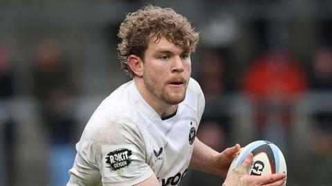 Ethan Staddon holds a rugby ball to his right in two hands as he runs on the pitch during a game last season. He is wearing a white Bath branded shirt