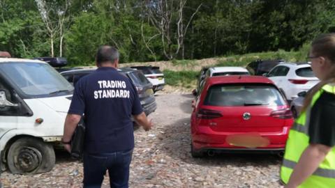Two trading standards officer inspecting vehicles parked next to a scrapyard.